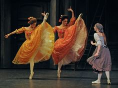 three ballerinas are performing on stage with their arms in the air and one is wearing an orange dress