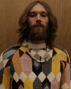 a man with long hair and a beard wearing a multicolored sweater stands in front of a wooden wall