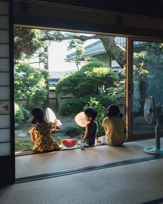 three children sitting on the windowsill looking out at an outside area with trees and bushes