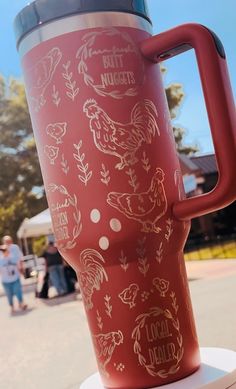 a red travel mug with roosters on it sitting on top of a white pedestal