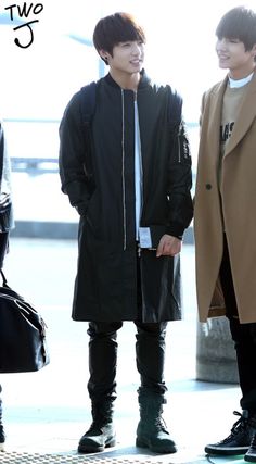 two young men standing next to each other on a sidewalk, one wearing a coat and the other in black