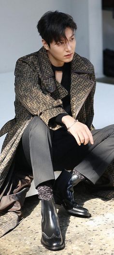 a young man sitting on the ground wearing black boots and a coat with an overcoat