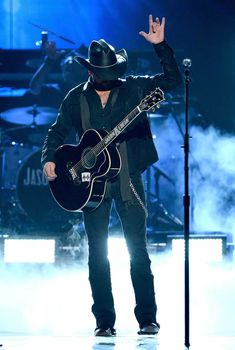 a man holding a guitar on stage with his hand in the air and wearing a cowboy hat