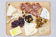 a wooden cutting board topped with cheese, meats and other foods next to olives