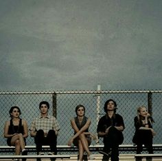 four people sitting on a bench in front of a chain link fence with the sky behind them