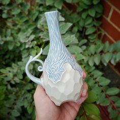 a hand holding a blue and white vase in front of some bushes with green leaves