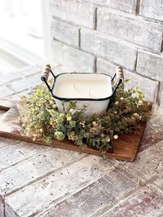 a candle is sitting on top of a wooden tray with flowers and greenery in front of a brick wall