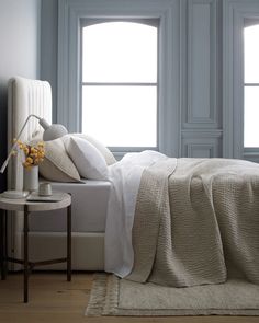 a white bed sitting next to two windows and a table with a vase on it