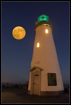 a white lighthouse with a green light on it's top and the moon in the background