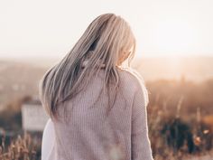 a woman with long blonde hair standing in tall grass looking at the sun behind her