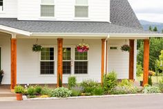 a white house with wooden pillars and flower pots on the front porch, surrounded by greenery