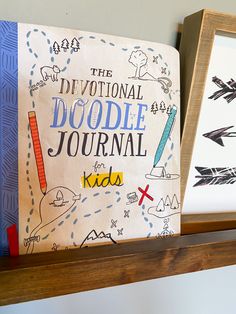 two children's drawings are on the shelf next to each other, and there is a book titled the national doodle journal for kids