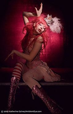 a woman with long red hair sitting on the ground in front of a dark background