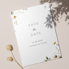 a white save the date card with daisies and leaves on it next to a dandelion