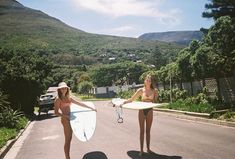 two women in bikinis holding surfboards on the side of a road with mountains in the background