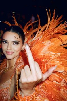 a woman in an orange feathered dress pointing at the camera with her finger up