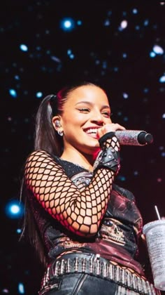 a woman in fishnet top holding a cup and microphone on stage with stars behind her