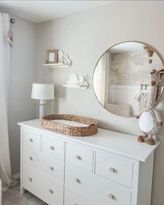 a white dresser with a basket on top and a round mirror above it in the corner