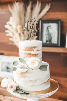 a three tiered cake with white flowers and greenery sits on a stand in front of a wooden wall