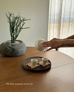 a person pouring water into two cups on top of a wooden table next to a potted plant