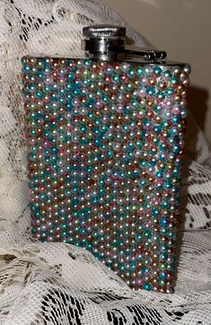 a flask covered in multicolored beads sits on a lace tablecloth next to a bottle opener