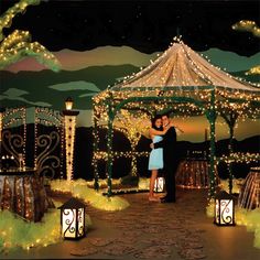 a man and woman standing under a gazebo covered in fairy lights at night with their arms around each other