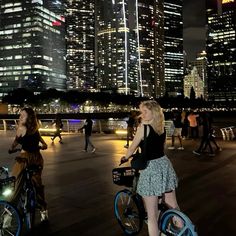 two women on bicycles in front of the city skyline at night, with skyscrapers lit up behind them