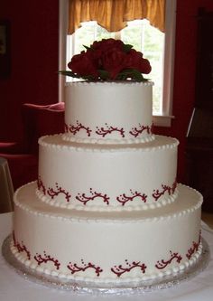 a three tiered wedding cake with red flowers on top and white frosting, sitting in front of a window