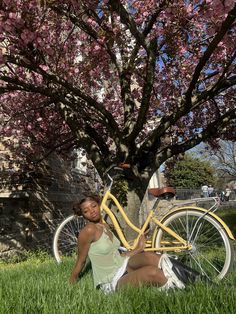 a woman sitting in the grass next to a bike under a tree with pink flowers