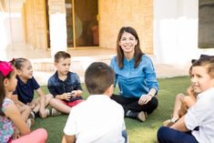 a woman sitting on the ground surrounded by small children in front of her, talking to each other