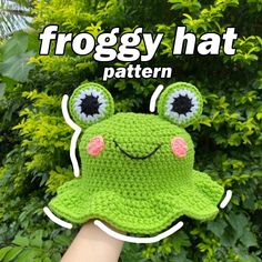 a crocheted froggy hat with eyes and ears is held up in front of some bushes