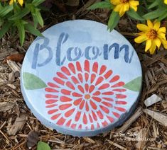 a painted rock with the word bloom on it in front of some yellow and red flowers