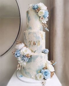 a three tiered wedding cake with blue and white flowers on the top, sitting on a table in front of a mirror