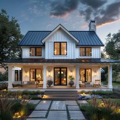 a large white house with lots of windows and lights on the front porch, surrounded by greenery