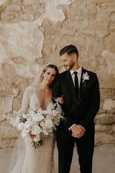 a bride and groom standing next to each other in front of a stone wall with white flowers