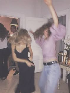 two young women are dancing in a room with other people and one is wearing a pink shirt