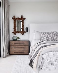 A modern bedroom with a wooden nightstand, decorative mirror, upholstered bed, patterned pillows, and a blanket. Neutral colors dominate the room, with a door leading outside. Nantucket Bedroom, Nantucket Family, Nantucket House, Workshop Apd, Family Compound, Architectural Ideas, Interior Minimalista, Fire Island