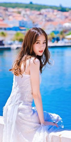 a woman in a white dress is sitting on a ledge by the water and looking at the camera