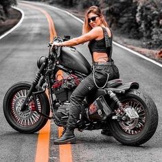 a woman sitting on top of a motorcycle in the middle of an empty road with trees and bushes behind her