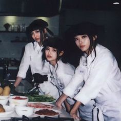 three women in white uniforms preparing food on a counter top with bowls and utensils