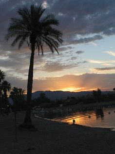 a palm tree on the beach at sunset with boats in the water and mountains in the background