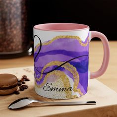 a purple and gold coffee mug sitting on top of a wooden table next to a cookie