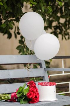 a white cake sitting on top of a wooden bench next to red flowers and balloons