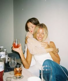 two women hugging each other while sitting at a table with drinks and wine glasses on it