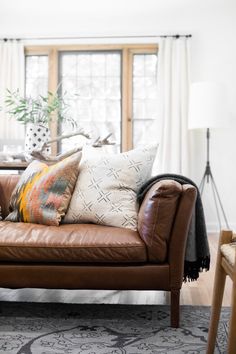 a brown leather couch sitting in front of a window with pillows on top of it