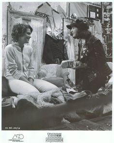 two women sitting on a bed talking to each other in a room full of clothes