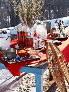 a picnic table with food and drinks on it in the snow, surrounded by trees