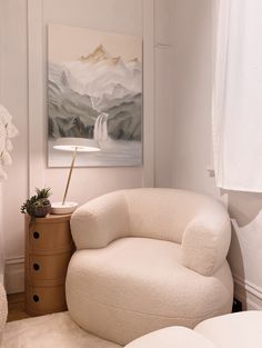 a white chair sitting in front of a painting on the wall next to a lamp