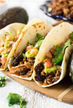 three tacos on a cutting board with avocado and cilantro in the background