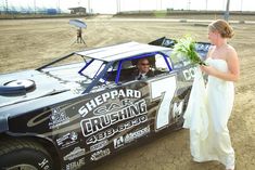 a woman in a wedding dress standing next to a race car with the driver looking at it